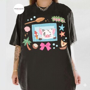 May include: Black graphic t-shirt featuring a cartoon illustration with a pig, chicken, and other tropical elements. The design includes a pink bow, starfish, and a small boat. The shirt has a relaxed fit and short sleeves.