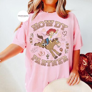 May include: A light pink short-sleeve t-shirt featuring a graphic of the character Jessie riding a horse. The text reads "HOWDY PARTNER" in a curved font. Additional illustrations include flowers, a cactus, a horseshoe, and a heart.