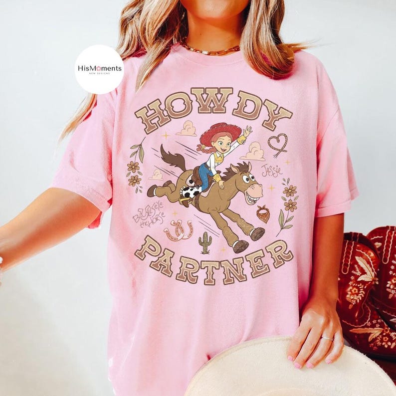 May include: A light pink short-sleeve t-shirt featuring a graphic of the character Jessie riding a horse. The text reads "HOWDY PARTNER" in a curved font. Additional illustrations include flowers, a cactus, a horseshoe, and a heart.