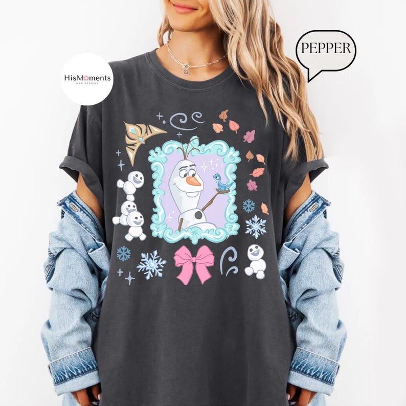 May include: Dark gray t-shirt featuring a colorful graphic of Olaf from Frozen, surrounded by snowflakes, a pink bow, and the text "PEPPER". The shirt has rolled-up sleeves and a relaxed fit.