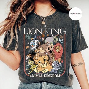 May include: Dark gray t-shirt featuring a colorful graphic of characters from Disney's The Lion King, including Simba, Timon, Pumbaa, and Mufasa. The text "THE LION KING ANIMAL KINGDOM" is displayed above and below the characters in a vintage style.