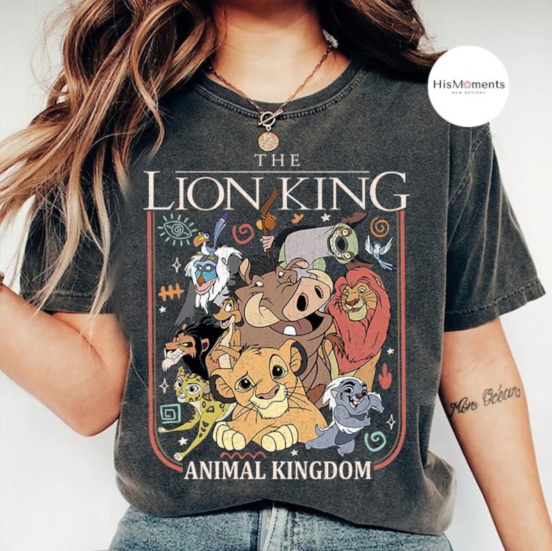 May include: Dark gray t-shirt featuring a colorful graphic of characters from Disney's The Lion King, including Simba, Timon, Pumbaa, and Mufasa. The text "THE LION KING ANIMAL KINGDOM" is displayed above and below the characters in a vintage style.