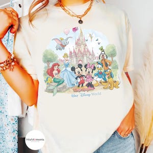 Pu&ograve; includere: Una t-shirt color crema con una grafica colorata di personaggi Disney tra cui Topolino, Minnie, Paperino, Pippo, Pluto, Ariel e Dumbo davanti a un castello. Il testo riporta Magic Kingdom e Walt Disney World.