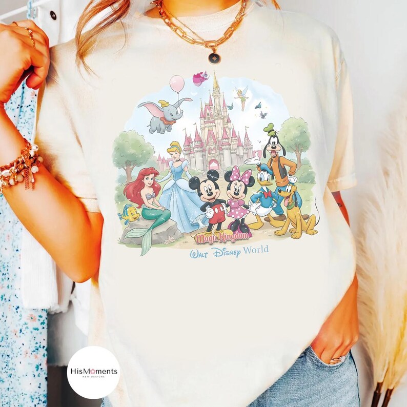 Pu&ograve; includere: Una t-shirt color crema con una grafica colorata di personaggi Disney tra cui Topolino, Minnie, Paperino, Pippo, Pluto, Ariel e Dumbo davanti a un castello. Il testo riporta Magic Kingdom e Walt Disney World.