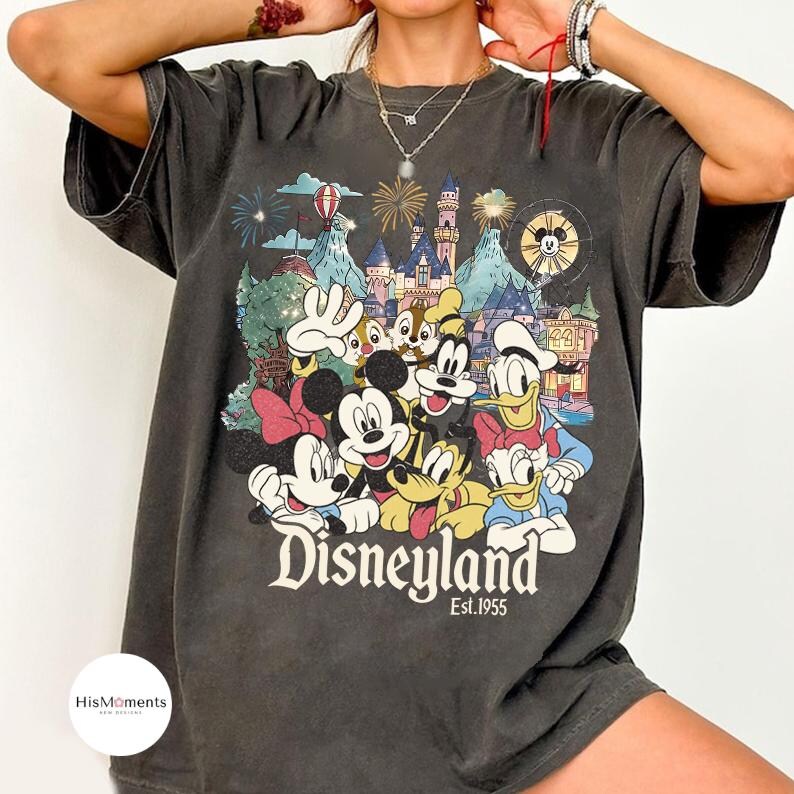 May include: A charcoal gray short-sleeve t-shirt featuring a colorful graphic print of various cartoon characters in front of a castle. The text Disneyland Est. 1955 is printed in white below the characters. The shirt has a crew neckline.