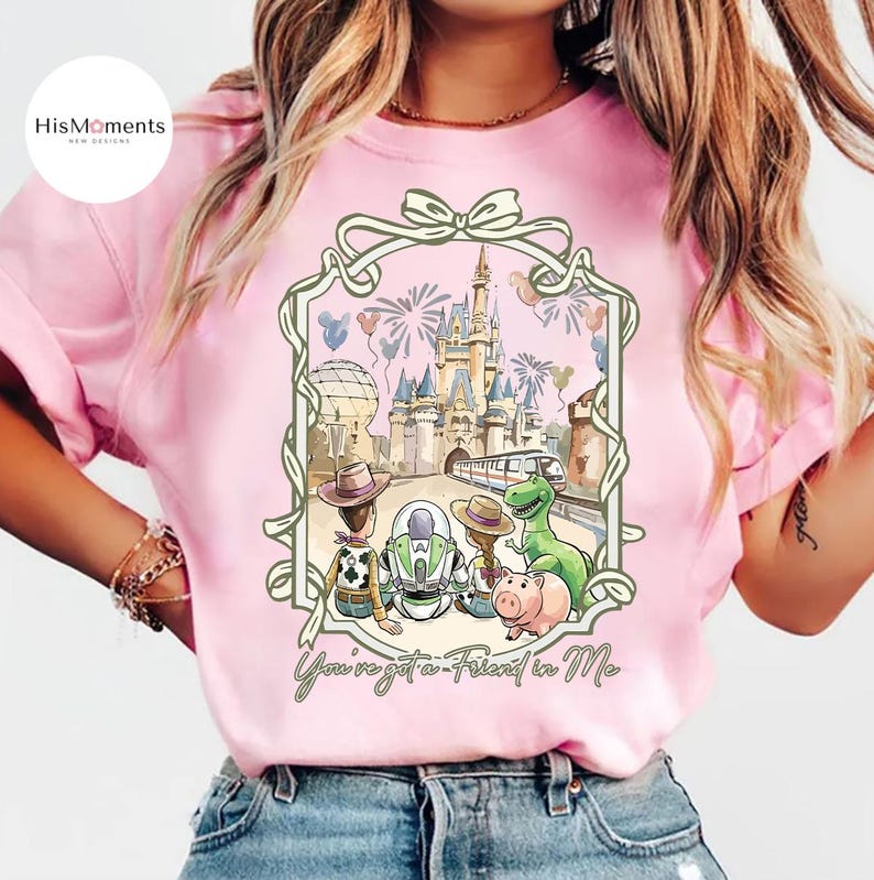 Peut inclure: T-shirt rose clair avec une illustration encadr&eacute;e des personnages de Toy Story devant un ch&acirc;teau, des feux d'artifice et un train. Le texte "You've got a Friend in Me" est en bas. Le t-shirt a des manches courtes.
