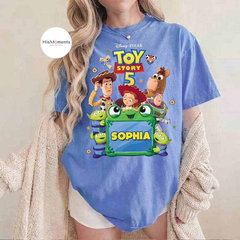 May include: A light blue t-shirt featuring a colorful graphic from the Disney Pixar movie Toy Story. The design includes characters like Woody, Buzz Lightyear, and others, with the name "Sophia" displayed prominently. The shirt is a comfortable, casual style.