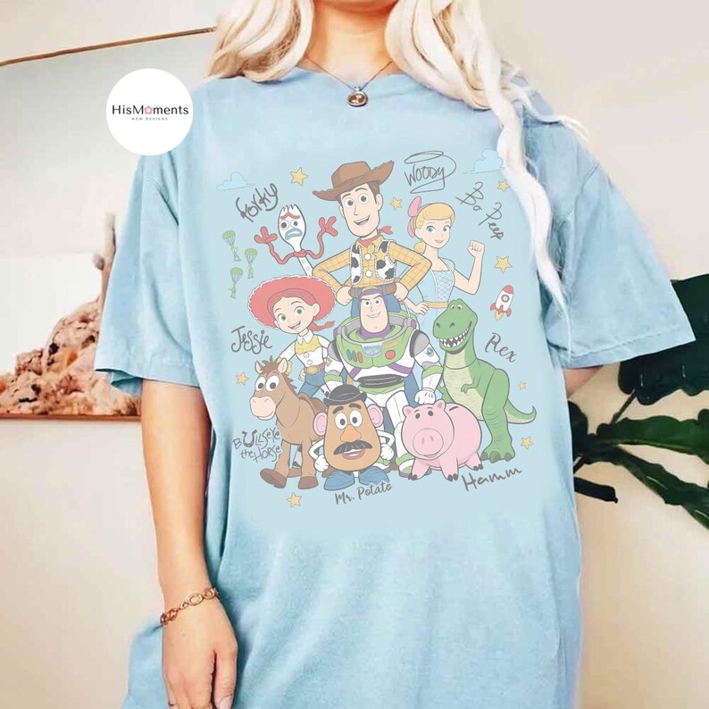 Peut inclure: Un t-shirt bleu clair avec une impression graphique repr&eacute;sentant divers personnages anim&eacute;s, dont un cow-boy, un ranger de l'espace, une cow-girl, un dinosaure, un cochon, une pomme de terre, un cheval et une fourchette, avec des noms manuscrits.