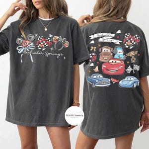 Może przedstawiać: Ciemnoszara koszulka z nadrukiem graficznym z napisem "Radiator Springs" i ilustracjami samochod&oacute;w. Projekt zawiera uszy Myszki Miki, wzory w szachownicę i kokardy. Tył koszulki r&oacute;wnież ma podobny wz&oacute;r z postaciami samochod&oacute;w.