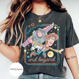 May include: Dark gray t-shirt featuring a colorful graphic of Buzz Lightyear and Woody from Toy Story, with the text "TO INFINITY and beyond" in a retro design. The shirt has a vintage, worn look.