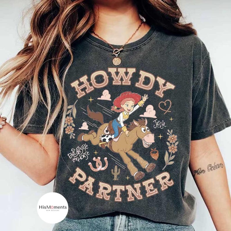 May include: A charcoal gray short-sleeve t-shirt featuring a graphic of the character Jessie riding Bullseye the horse. The text reads HOWDY PARTNER and Jessie. Decorative elements include flowers, clouds, stars, a heart, and a horseshoe.