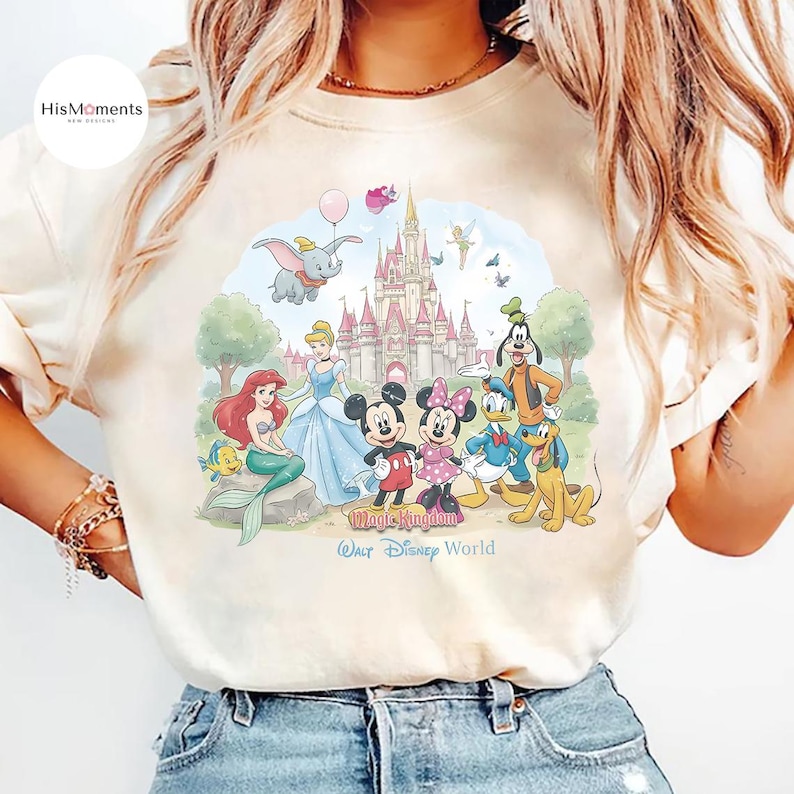 Pu&ograve; includere: Una t-shirt color crema con una grafica colorata di personaggi Disney tra cui Topolino, Minnie, Ariel, Cenerentola, Paperino, Pippo, Pluto e Dumbo davanti a un castello con la scritta Walt Disney World.
