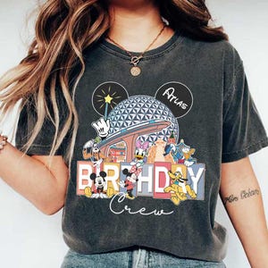May include: A grey t-shirt featuring a graphic of Mickey Mouse and other Disney characters in front of the Spaceship Earth ride at Epcot. The text "Birthday Crew" is printed below the characters. The text "Atlas" is written on the top right corner of the shirt.