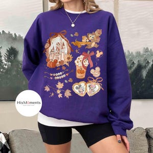 Op de afbeelding: Een paarse sweatshirt met Disney-thema herfstafbeeldingen. Het ontwerp bevat een kasteel, Knabbel en Babbel, Mickey en Minnie Mouse, een pumpkin spice latte en herfstbladeren. De tekst "HisMoments new designs" staat op de sweatshirt.