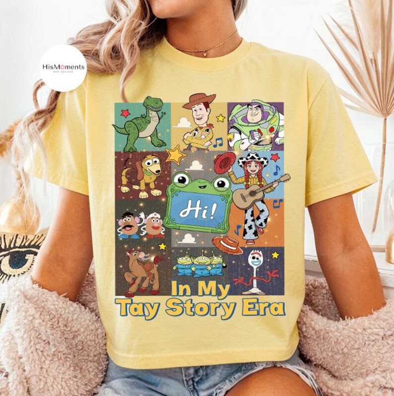 May include: A light yellow t-shirt featuring a graphic print of various Toy Story characters including Rex, Woody, Buzz Lightyear, Slinky Dog, Mr. and Mrs. Potato Head, Jessie, and Forky. The text reads "Hi!" and "In My Tay Story Era" in blue.