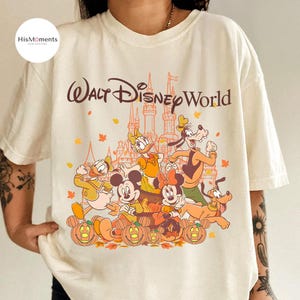 Op de afbeelding: Cr&egrave;mekleurig T-shirt met een Walt Disney World-afbeelding. Het ontwerp bevat Mickey Mouse, Minnie Mouse, Donald Duck, Daisy Duck, Goofy en Pluto, met een kasteel en pompoenen. De tekst "Walt Disney World" staat bovenaan gebogen.
