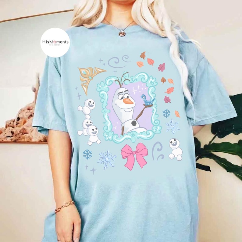 May include: Light blue t-shirt featuring a framed illustration of Olaf from Frozen, with a blue bird, snowflakes, and other winter-themed graphics. The shirt also includes the text "HisMoments" in a white circle.