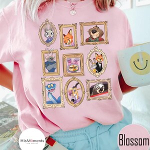 Puede incluir: Camiseta rosa claro con retratos enmarcados en oro de personajes de dibujos animados. La camiseta tiene la palabra "Blossom" impresa. La marca "HisMoments" tambi&eacute;n est&aacute; impresa en la camiseta. La camiseta es de corte holgado.