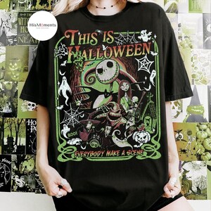 K&ouml;nnte beinhalten: Schwarzes T-Shirt mit einer farbenfrohen Grafik aus "The Nightmare Before Christmas". Das Design zeigt Jack Skellington, Sally und andere Charaktere mit dem Text "THIS IS HALLOWEEN" und "EVERYBODY MAKE A SCENE" in Gr&uuml;n und Rot.