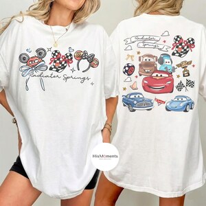 Może przedstawiać: Biała koszulka z kolorową grafiką postaci z filmu Auta Disneya i napisem "Radiator Springs". Projekt zawiera obrazy Zygzaka McQueena, Złomka i innych postaci, a także uszy Myszki Miki i szachownice.