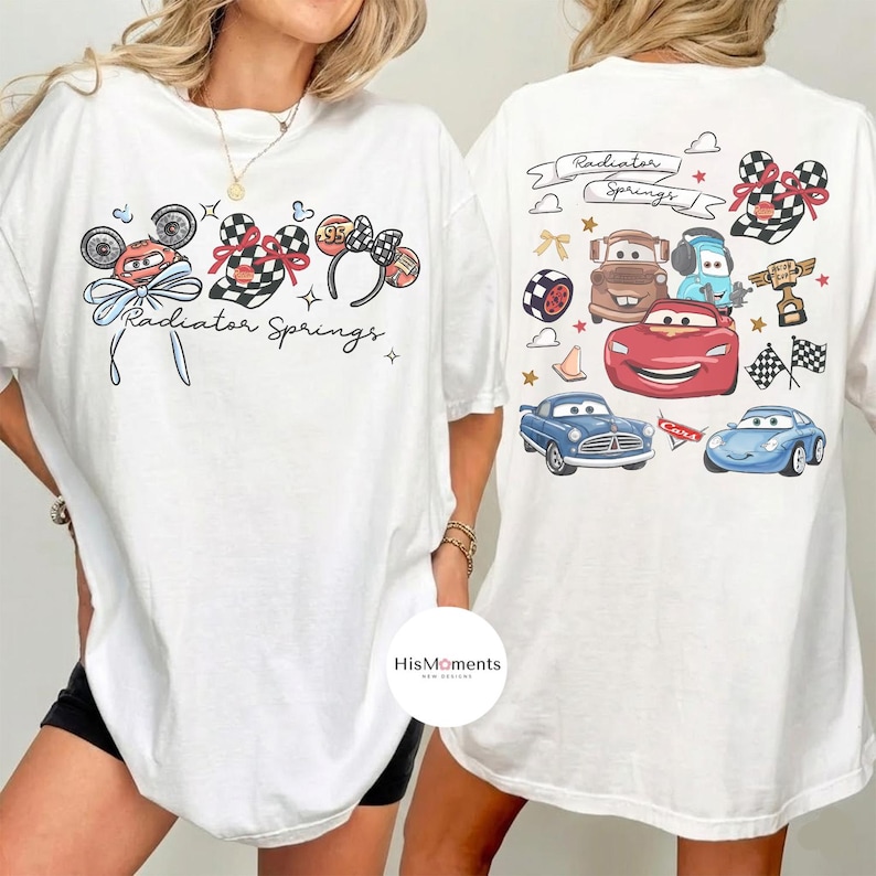 Może przedstawiać: Biała koszulka z kolorową grafiką postaci z filmu Auta Disneya i napisem "Radiator Springs". Projekt zawiera obrazy Zygzaka McQueena, Złomka i innych postaci, a także uszy Myszki Miki i szachownice.