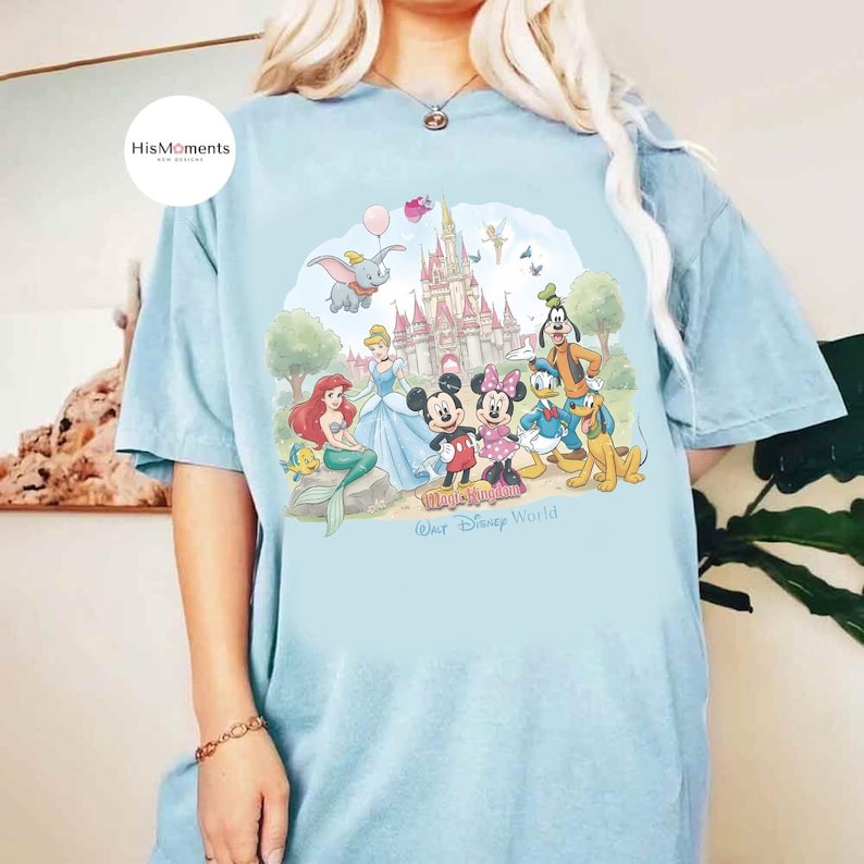 Pu&ograve; includere: Una t-shirt azzurra con una grafica colorata di personaggi Disney tra cui Topolino, Minnie, Paperino, Pippo, Pluto, Ariel, Cenerentola e Dumbo davanti a un castello con la scritta Magic Kingdom Walt Disney World.