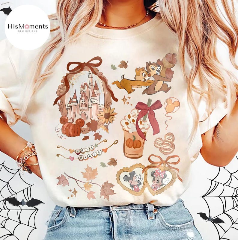 Op de afbeelding: Cr&egrave;mekleurig T-shirt met herfstthema-afbeeldingen. Het ontwerp omvat een kasteel, pompoenen, Chip en Dale, een pumpkin spice latte en Mickey en Minnie Mouse. De woorden "Fall Vibes" zijn ook aanwezig. Het shirt heeft een ronde hals en korte mouwen.