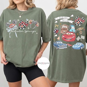Może przedstawiać: Oliwkowo-zielona koszulka z motywem z filmu Auta. Prz&oacute;d koszulki przedstawia napis "Radiator Springs" z grafiką samochod&oacute;w. Tył koszulki r&oacute;wnież ma wz&oacute;r "Radiator Springs" z r&oacute;żnymi postaciami z samochod&oacute;w.