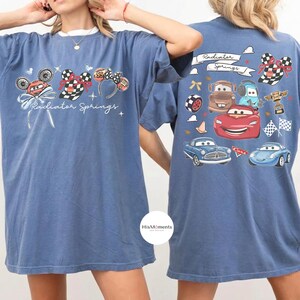 Może przedstawiać: Niebieskie koszulki z grafiką przedstawiającą postacie i elementy z filmu "Auta". Prz&oacute;d koszulki przedstawia uszy Myszki Miki, szachownicowe kokardy i napis "Radiator Springs". Tył przedstawia r&oacute;żne ilustracje samochod&oacute;w i napis "Radiator Springs."