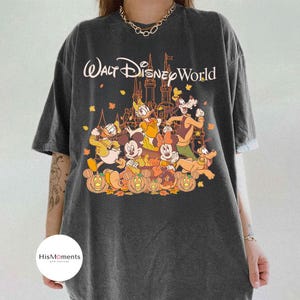 Op de afbeelding: Donkergrijze t-shirt met een afbeelding van Disney-personages voor een kasteel en de tekst "Walt Disney World". Het ontwerp bevat Mickey Mouse, Donald Duck, Goofy en pompoenen.