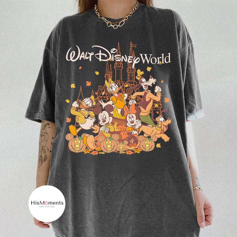 Op de afbeelding: Donkergrijze t-shirt met een afbeelding van Disney-personages voor een kasteel en de tekst "Walt Disney World". Het ontwerp bevat Mickey Mouse, Donald Duck, Goofy en pompoenen.