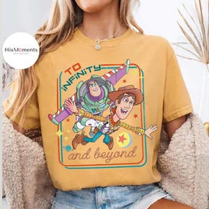 May include: Mustard yellow t-shirt featuring a graphic of Buzz Lightyear and Woody from Toy Story. The design includes the text "TO INFINITY and beyond" in a retro style. The shirt has short sleeves and a crew neck.