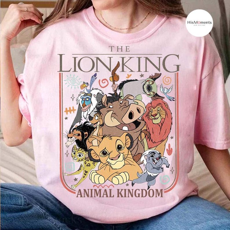 May include: A light pink t-shirt featuring a colorful graphic of characters from The Lion King. The design includes Simba, Timon, Pumbaa, and other characters, with the words "THE LION KING" and "ANIMAL KINGDOM" in a rectangular frame.