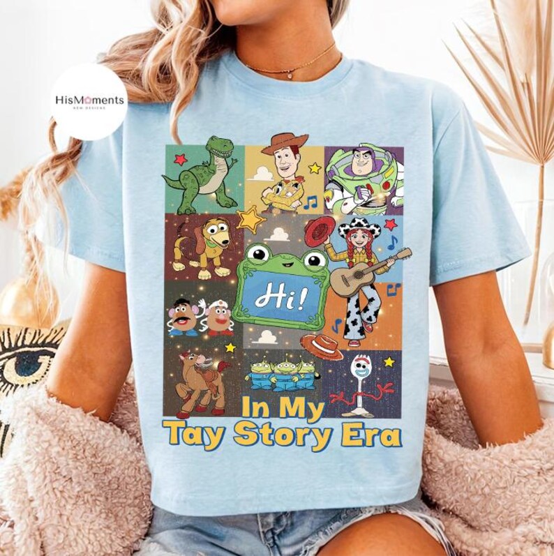 May include: A light blue t-shirt featuring a graphic print of various animated characters from the Toy Story franchise, including a green dinosaur, a cowboy, a space ranger, and a cowgirl playing a guitar, with the text "Hi!" and "In My Tay Story Era."