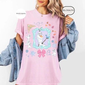 May include: A light pink t-shirt featuring a cartoon image of Olaf from Frozen, surrounded by snowflakes, leaves, and a pink bow. The shirt has the text "HisMoments" on the upper left and "BLOSSOM" on the upper right.
