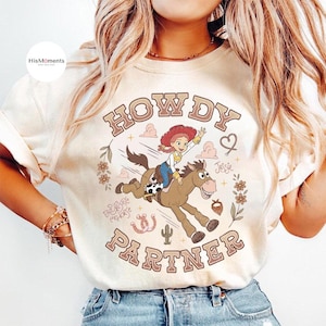 May include: A cream-colored t-shirt featuring a graphic of the character Jessie riding Bullseye the horse. The text reads HOWDY PARTNER, Jessie, and Bullseye the horse. The design includes floral elements, a horseshoe, a cactus, and a heart.