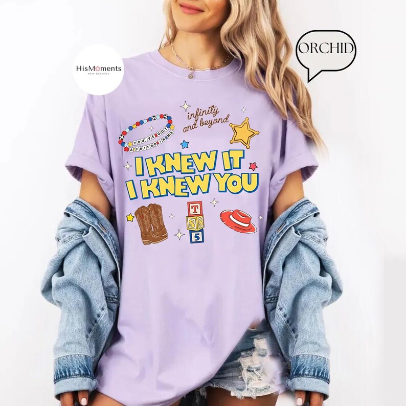 K&ouml;nnte beinhalten: Ein hellviolettes T-Shirt mit dem Text I KNEW IT I KNEW YOU in Gelb mit blauen Umrandungen. Die Grafiken zeigen ein Perlenarmband, einen gelben Stern, einen braunen Cowboystiefel, Alphabet-W&uuml;rfel mit T, S, 5 und einen roten Cowboyhut.