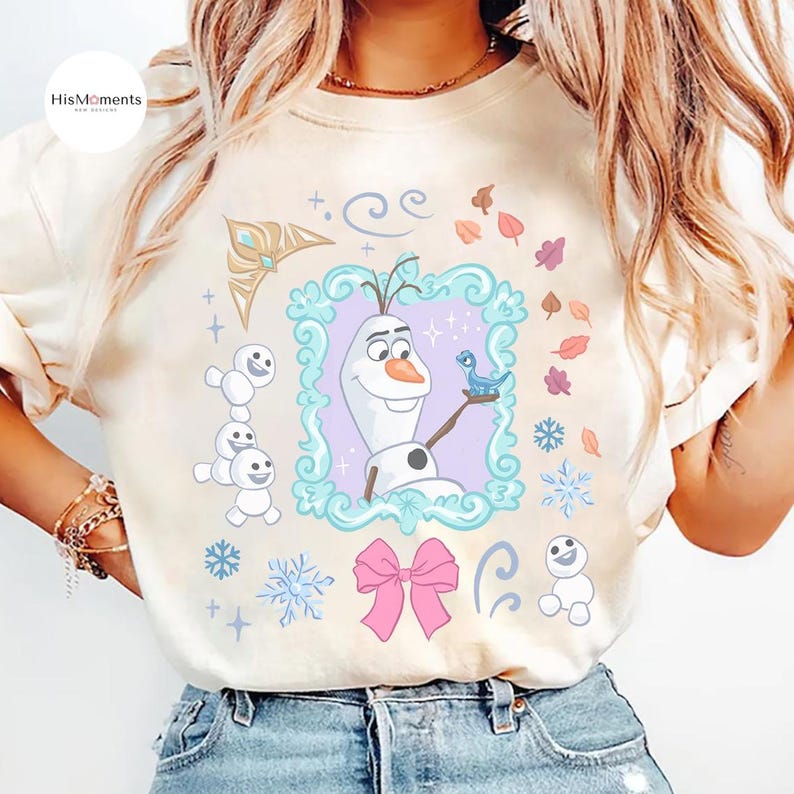 May include: Cream-colored t-shirt featuring a colorful graphic of Olaf from Frozen. Olaf is in a decorative frame, holding a blue bird. The design includes snowflakes, a pink bow, and other winter-themed elements. The shirt has a slightly faded, vintage look.