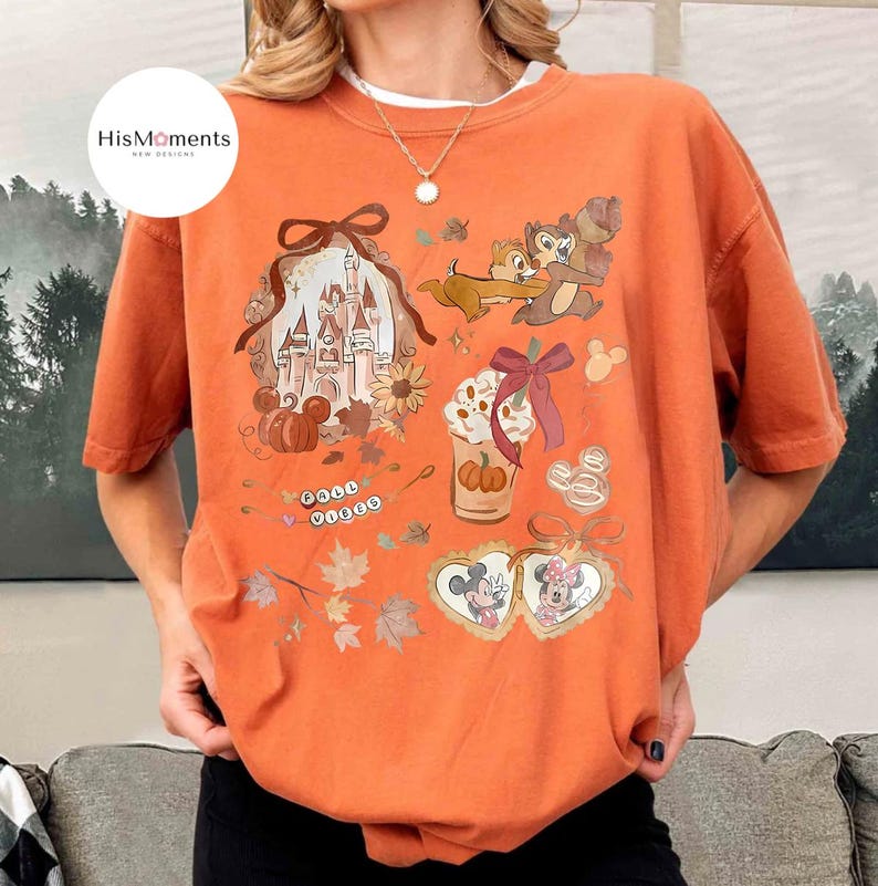 Op de afbeelding: Oranje grafisch T-shirt met een Disney-thema herfstontwerp. De print bevat Chip en Dale, een kasteel, pompoenen en een koffiekopje. De tekst "Fall Vibes" is ook aanwezig.