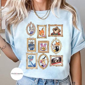 Puede incluir: Camiseta azul claro con retratos enmarcados de personajes de animales animados. Los retratos est&aacute;n en marcos dorados ornamentados e incluyen una variedad de animales como un conejo, un zorro y un perezoso. La camiseta tiene un logotipo blanco redondo que dice "HisMoments NEW DESIGNS".