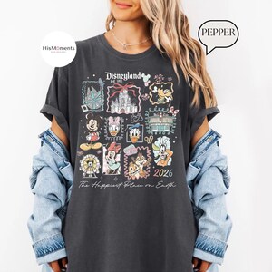 Peut inclure: T-shirt gris fonc&eacute; avec un motif color&eacute; de Disneyland, repr&eacute;sentant des personnages de dessins anim&eacute;s et des ic&ocirc;nes du parc. Le t-shirt affiche le texte "Disneyland Est. 1955" et "2026", ainsi que la phrase "The Happiest Place on Earth".