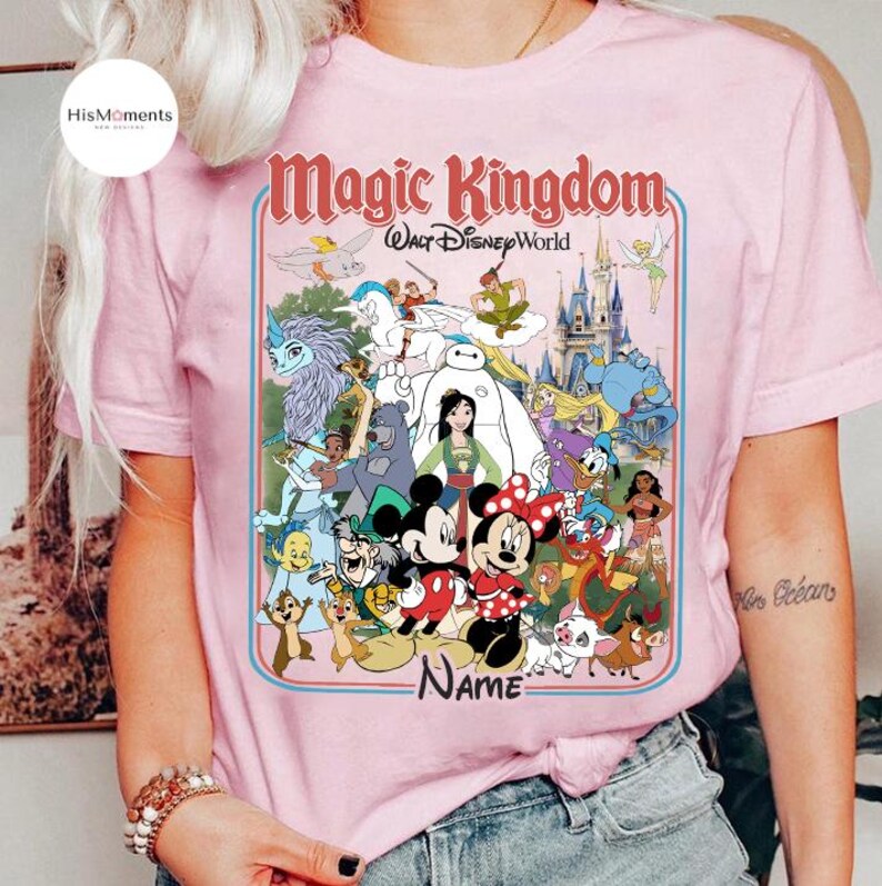 Puede incluir: Camiseta rosa claro con un gr&aacute;fico colorido de personajes de Disney, incluyendo Mickey y Minnie Mouse, con el texto "Magic Kingdom Walt Disney World". El dise&ntilde;o est&aacute; enmarcado en un borde azul claro.