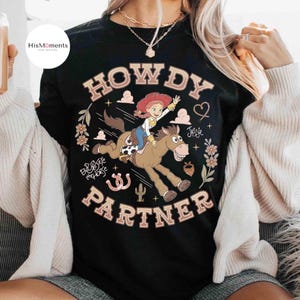 May include: A black t-shirt featuring a graphic print of a cowgirl riding a brown horse. The text reads HOWDY PARTNER, Jessie, and Bullseye the Horse. The design includes illustrations of a cactus, horseshoes, flowers, and clouds in shades of pink, white, and brown.