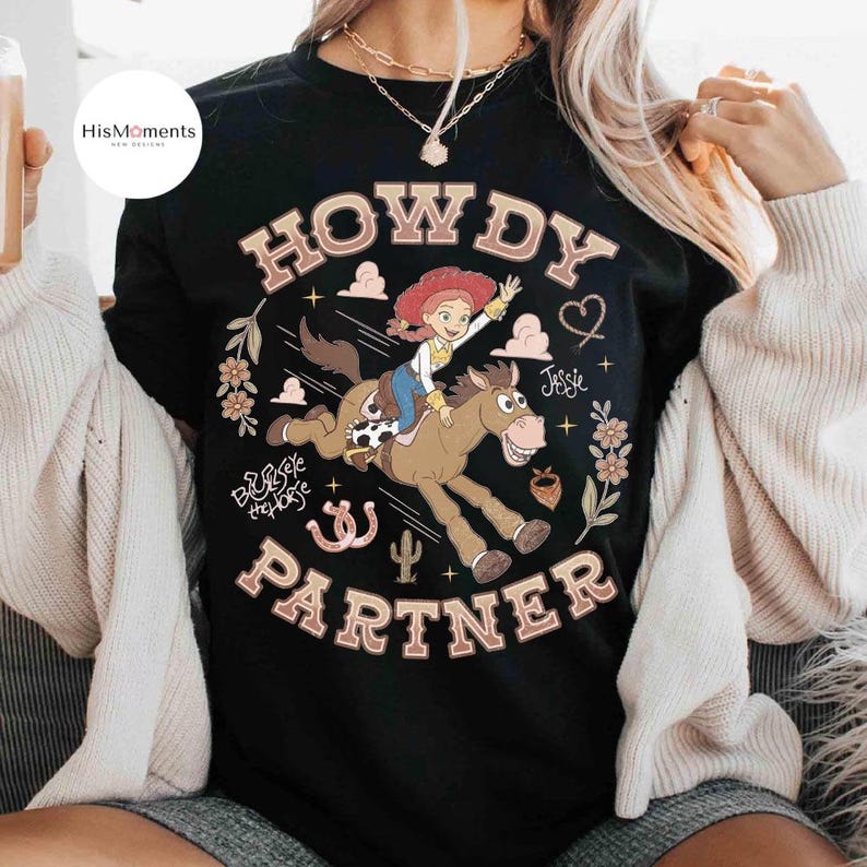 May include: A black t-shirt featuring a graphic print of a cowgirl riding a brown horse. The text reads HOWDY PARTNER, Jessie, and Bullseye the Horse. The design includes illustrations of a cactus, horseshoes, flowers, and clouds in shades of pink, white, and brown.