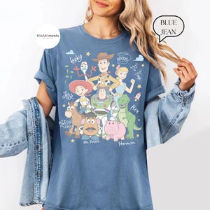 Peut inclure: Un t-shirt bleu jean avec une impression graphique des personnages de Toy Story, incluant Woody, Buzz l'&Eacute;clair, Jessie, Rex, Bayonne, Monsieur Patate, Fourchette, la Berg&egrave;re et Pile-Poil, avec des noms manuscrits et des &eacute;toiles.