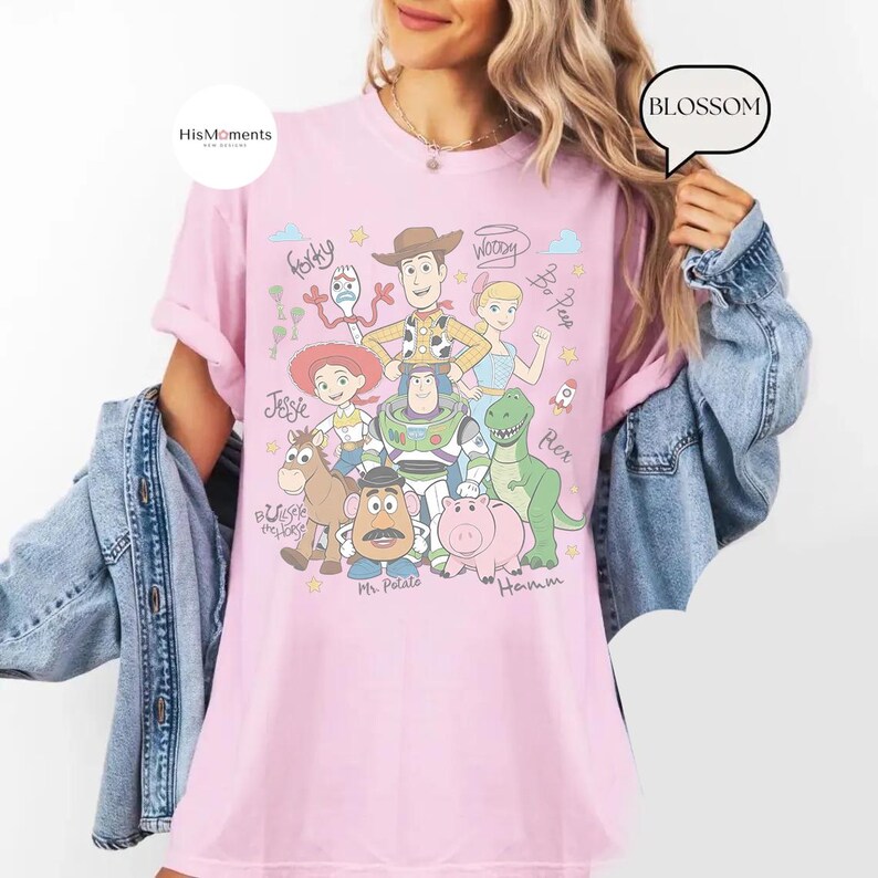 Peut inclure: Un t-shirt rose clair avec une impression graphique des personnages de Toy Story, notamment Woody, Buzz l'&Eacute;clair, Jessie, Rex, Hamm, Monsieur Patate, Fourchette, la Berg&egrave;re et Pile-Poil, accompagn&eacute;s de noms &eacute;crits &agrave; la main et d'&eacute;toiles.