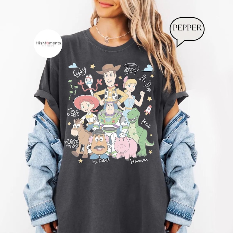 Peut inclure: Un t-shirt gris anthracite avec une impression graphique des personnages de Toy Story, dont Woody, Buzz l'&Eacute;clair, Jessie, Rex, Bayonne, Monsieur Patate, Pile-Poil, Fourchette et la Berg&egrave;re, avec leurs noms &eacute;crits en cursive et des &eacute;toiles.