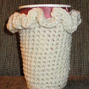 May include: White crocheted cup cozy with a ruffled top. The cozy is made with a simple stitch pattern and is designed to fit a standard-sized cup.