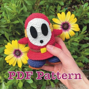 Könnte beinhalten: Gehäkelte Shy Guy-Puppe in Rot, Weiß, Schwarz, Gelb und Blau. Die Puppe hat eine weiße Maske mit schwarzen Augenlöchern und einem schwarzen Mund. Der Text "PDF Pattern" wird unten im Bild in Rosa angezeigt.