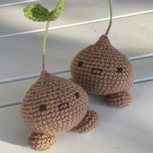 Peut inclure: Deux figurines amigurumi crochetées marron avec des corps arrondis et de petits pieds. Chaque figurine présente un sommet pointu, un visage brodé simple avec deux yeux et une bouche, et une fine tige verte avec une petite feuille verte au sommet.